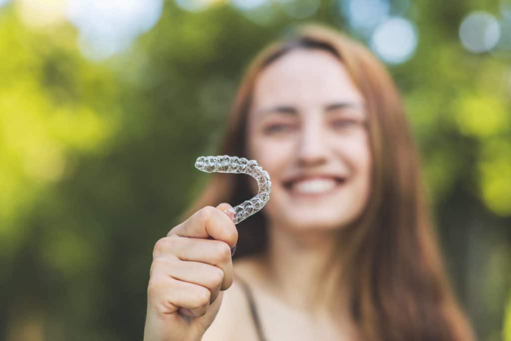 a young woman standing outside next to trees and holding invisalign aligners in one hand