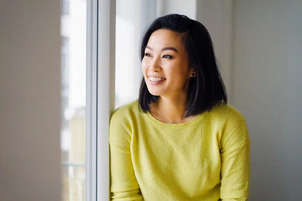 A smiling asian woman leaning against a window.