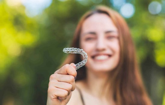 a young woman standing outside next to trees and holding invisalign aligners in one hand